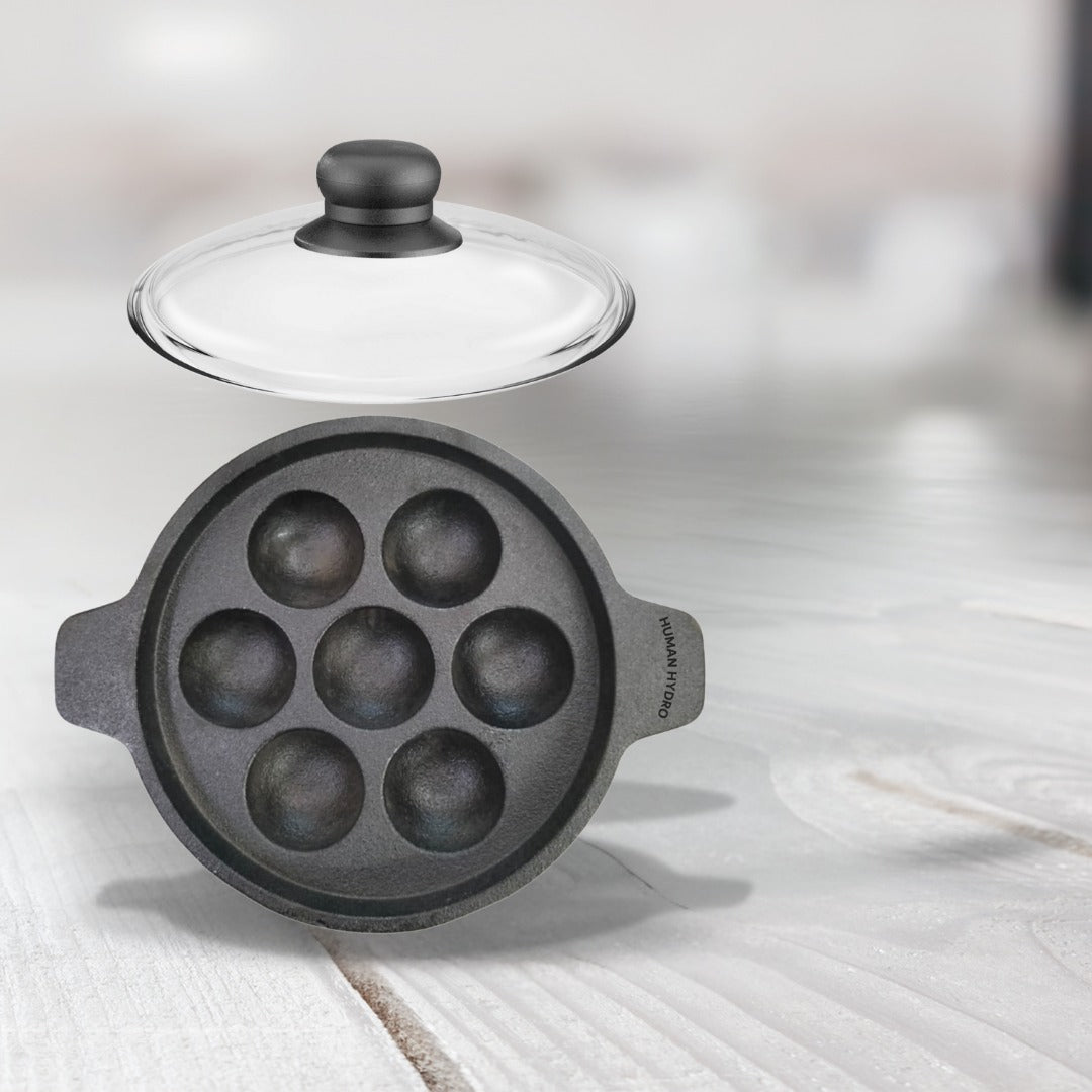 Cast iron paniyaram pan with a glass lid, designed for making traditional Indian paniyaram, featuring seven round cavities.