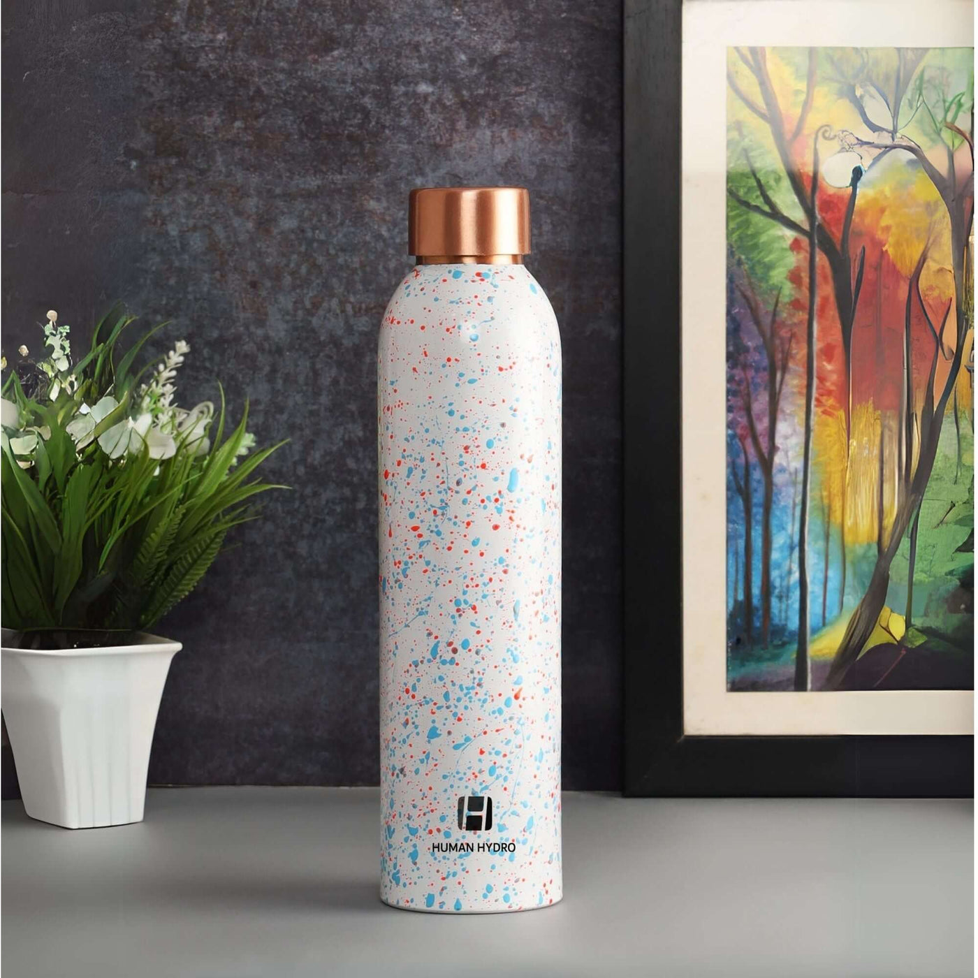 Stylish water bottle with speckled design and copper lid, placed next to a plant and vibrant artwork.